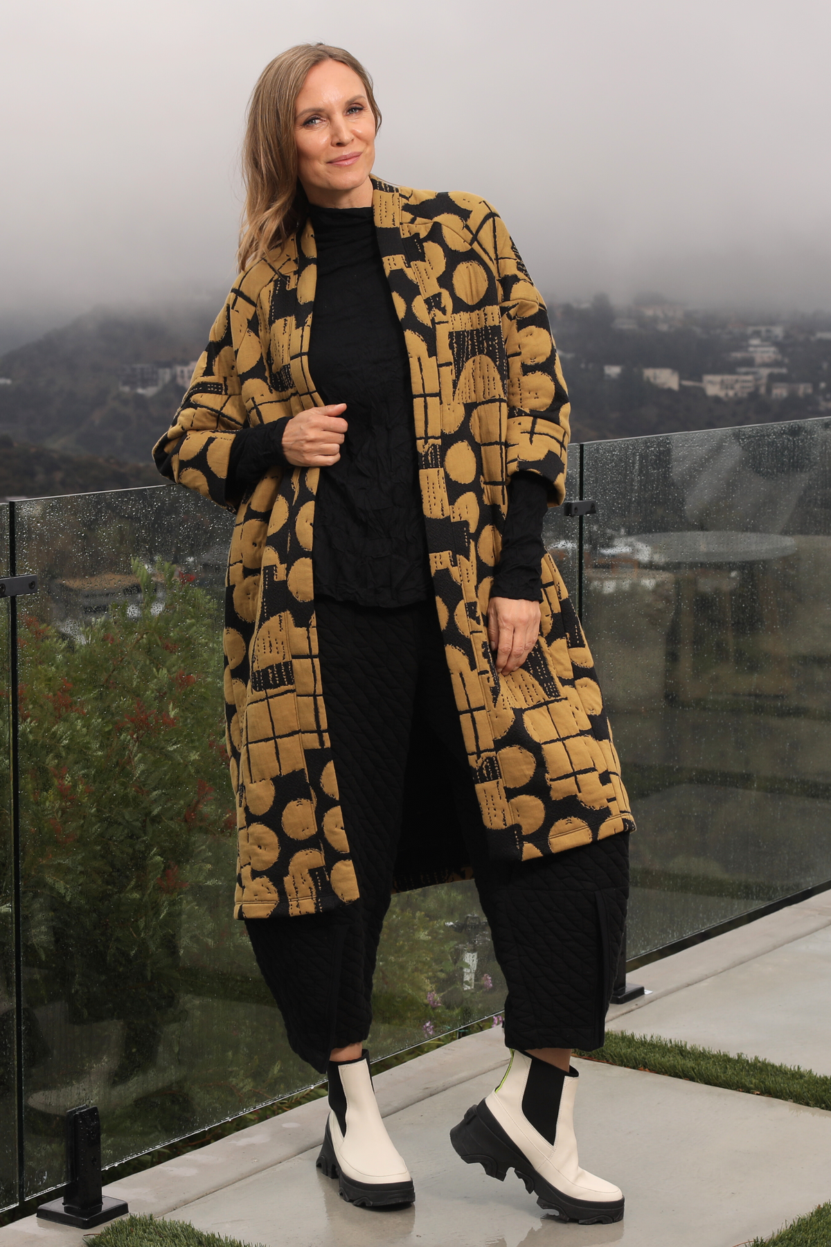Woman wearing a patterned coat on a balcony with a cityscape view.