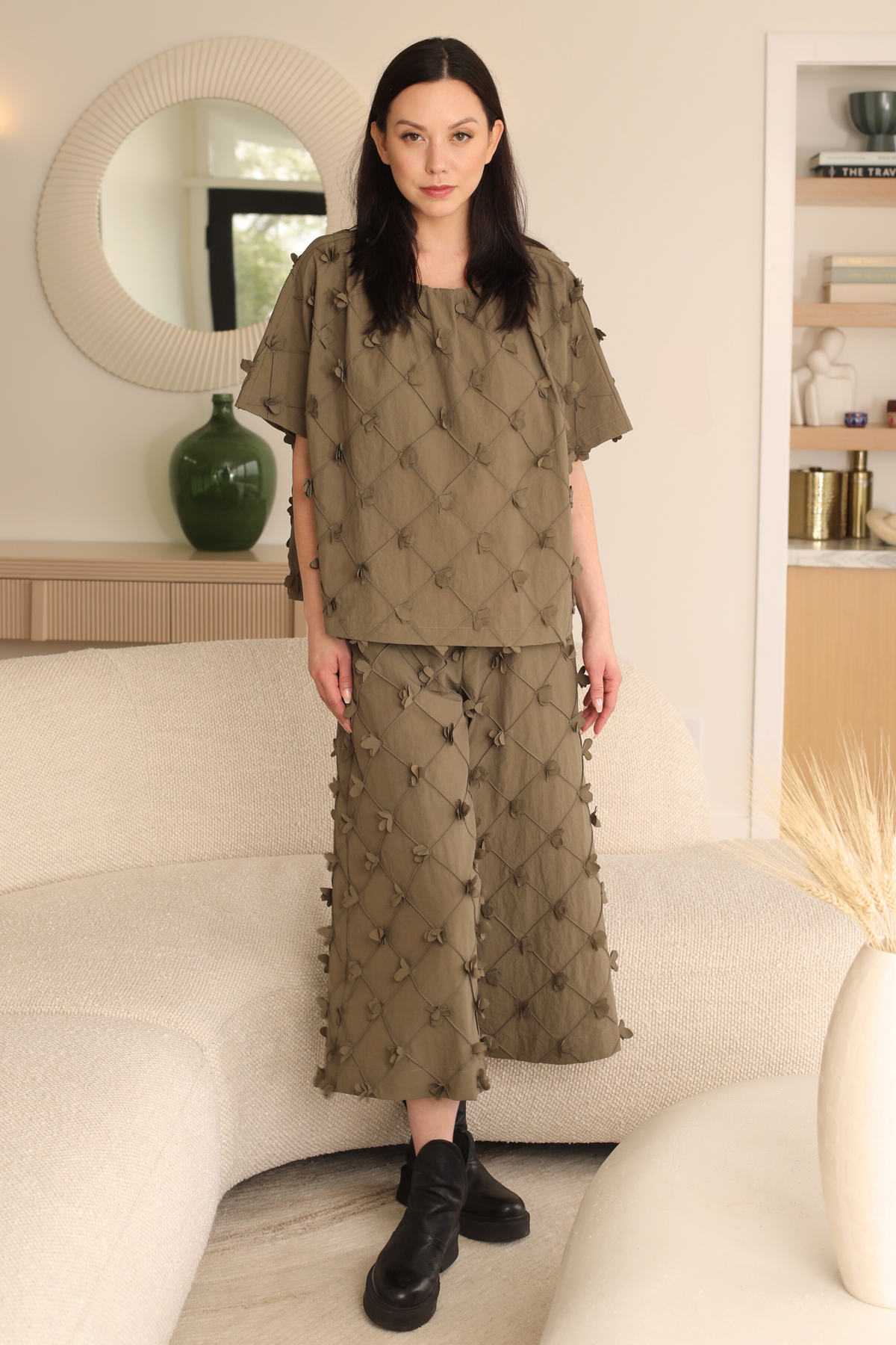 Woman wearing a green embroidered outfit in a room with a mirror and shelves.