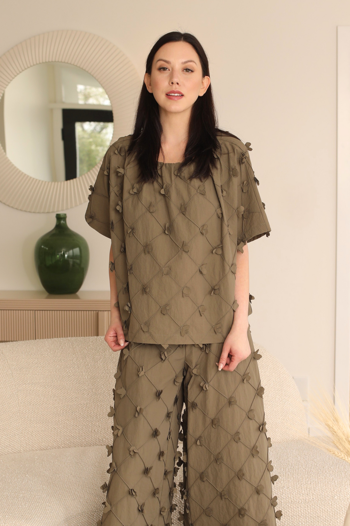 Woman wearing a green embroidered outfit in a room with a mirror and vase.