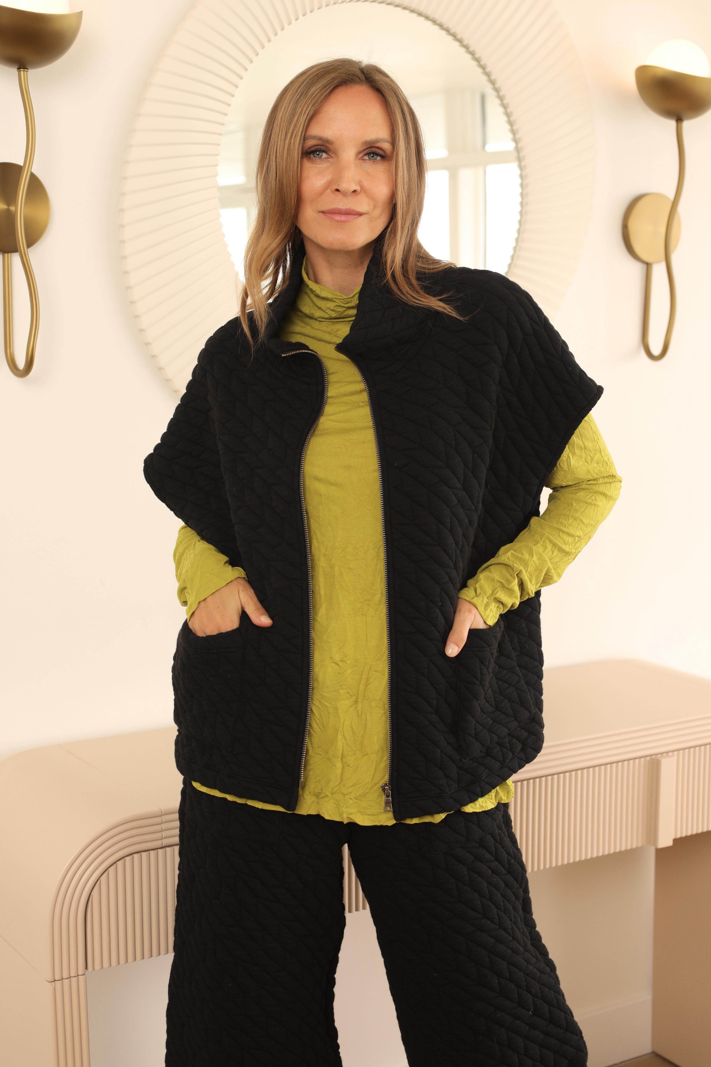 Woman wearing a black quilted vest over a green top and black pants in an indoor setting.