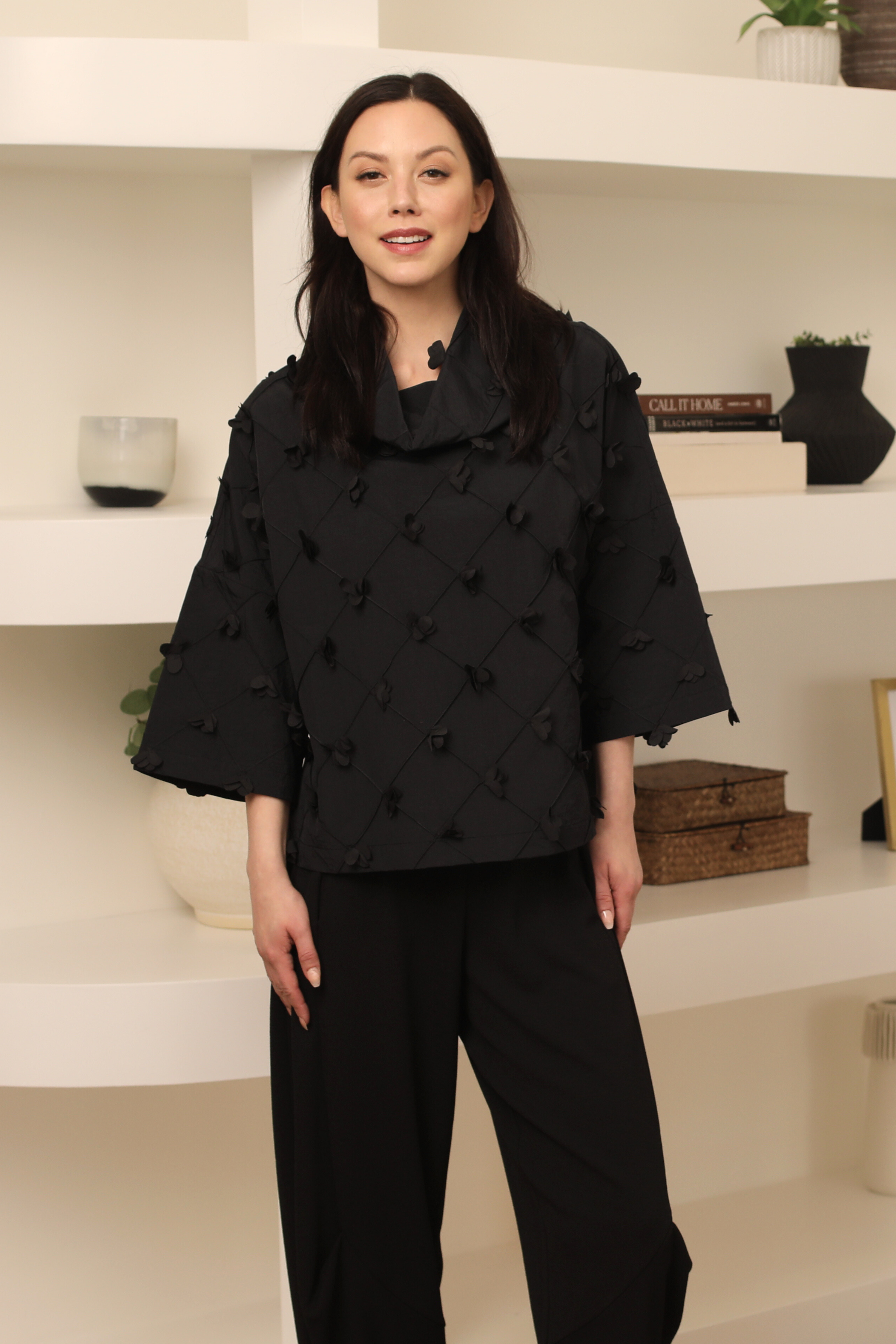Woman wearing a black textured outfit standing in front of a white shelf.