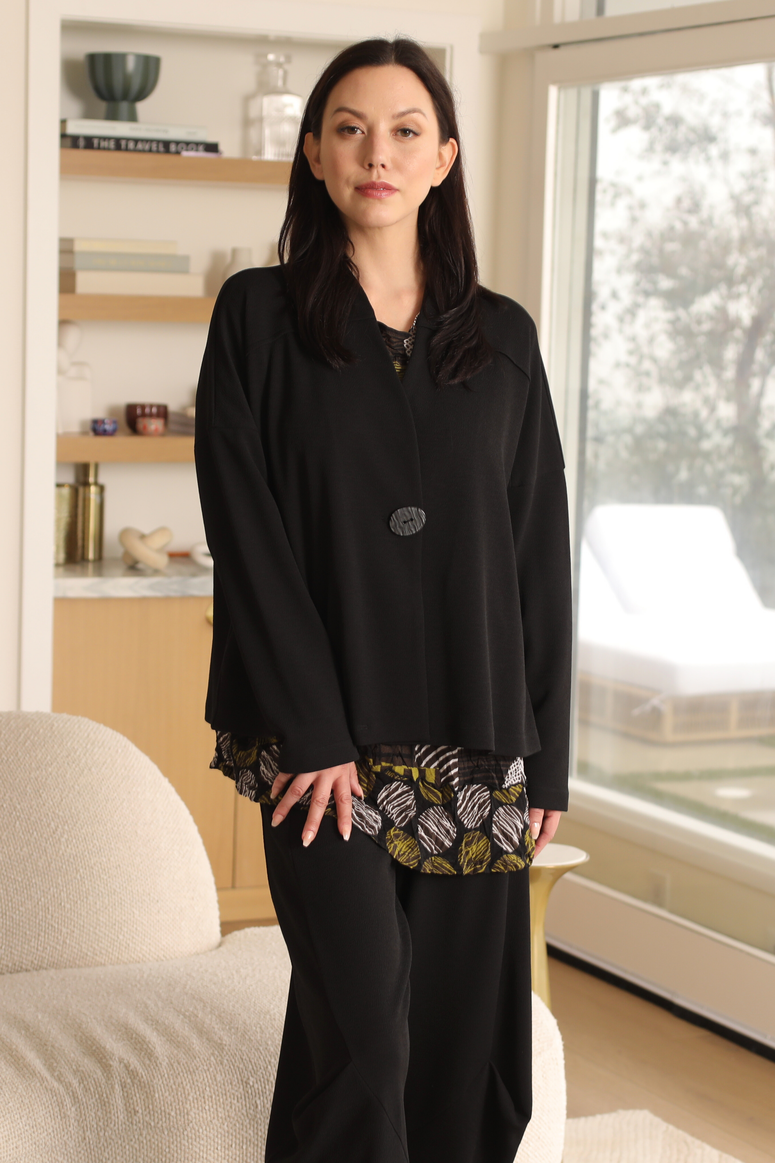 Woman wearing a black outfit with patterned layers in a room with a window and furniture.