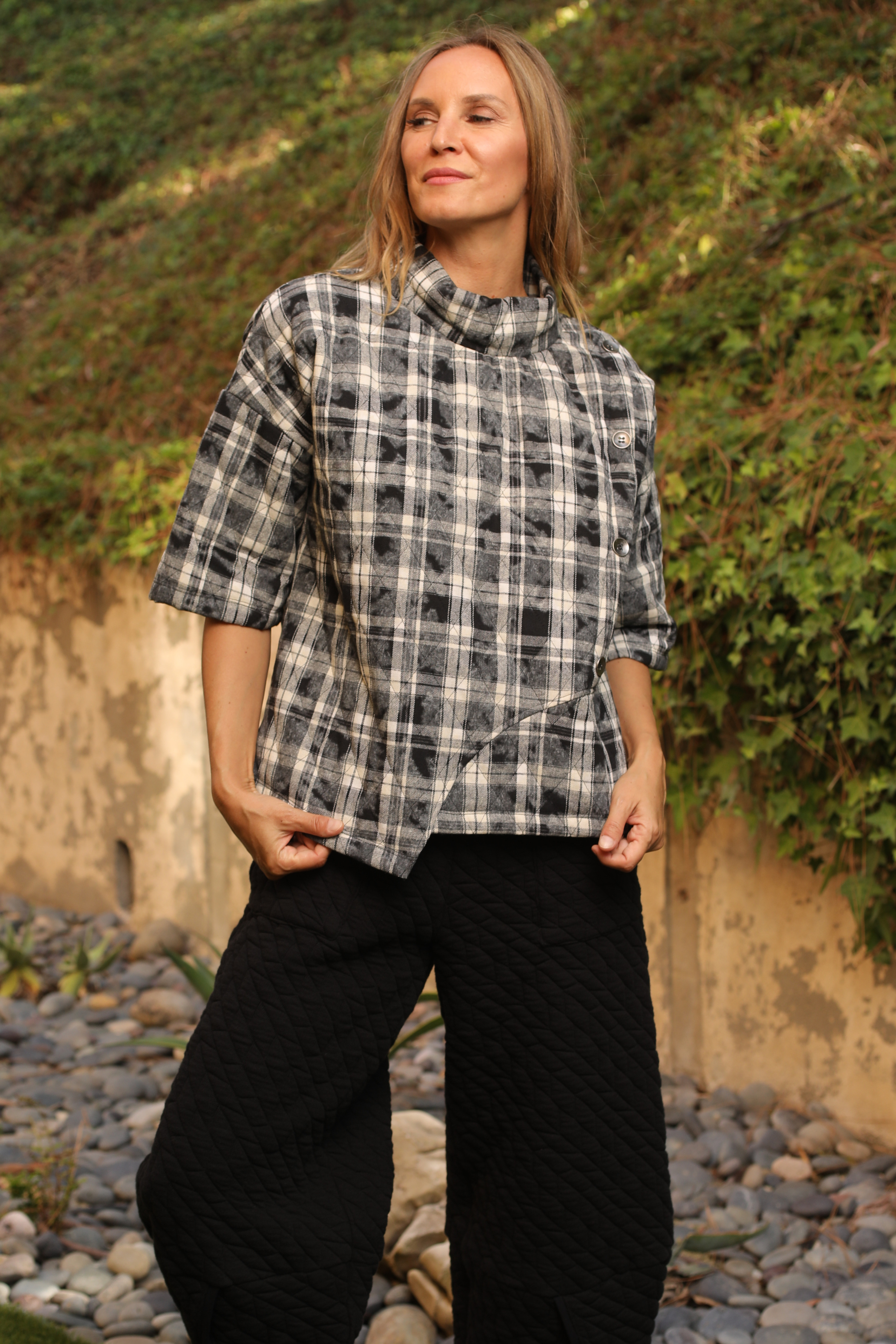 Woman wearing a plaid shirt and black pants standing outdoors with greenery in the background